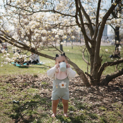 Bébé buvant dans un gobelet sous un arbre en fleurs, vêtu d'un body floral pour maternité et bien-être